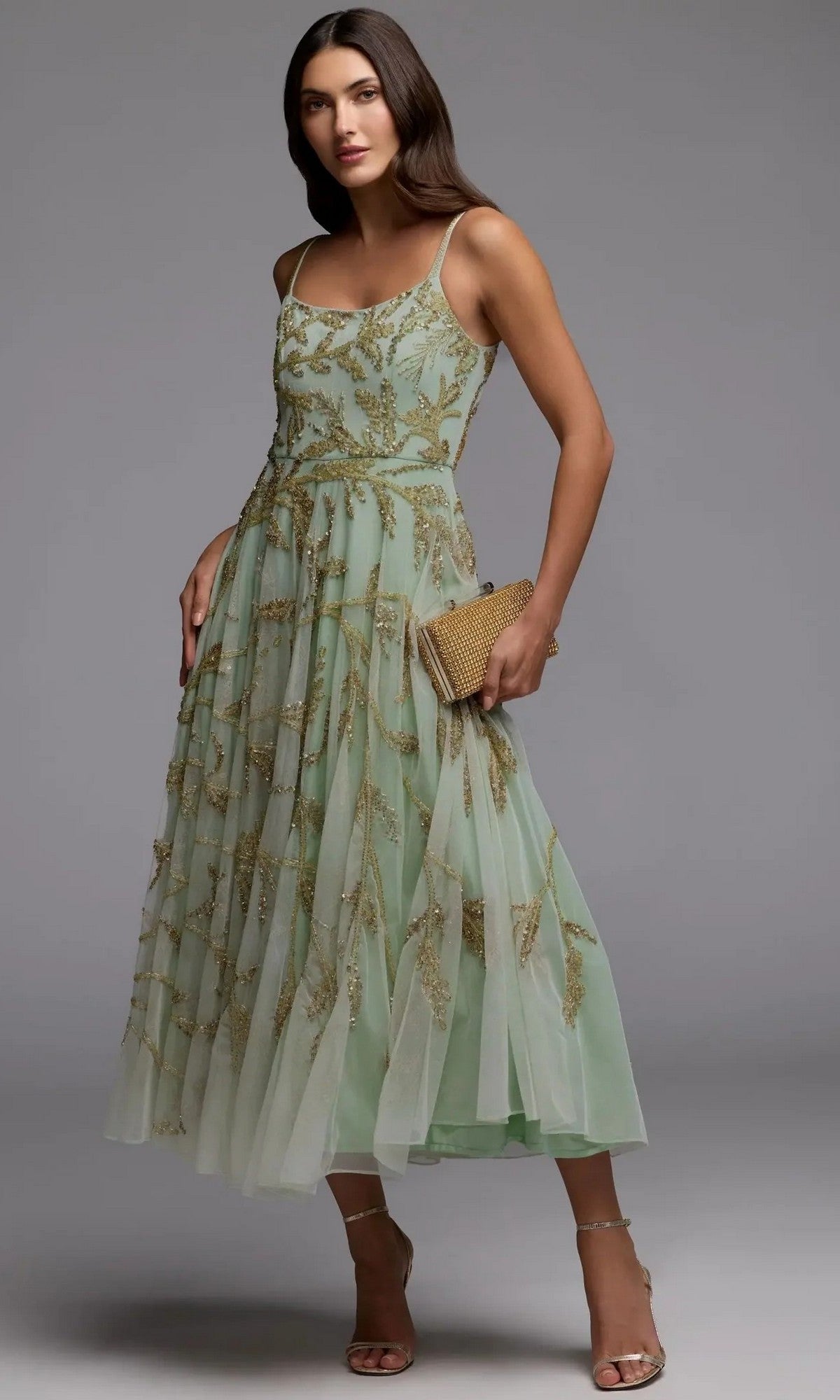 A woman wearing the Mac Duggal 6255 tea-length party dress in seafoam gold, featuring gold beaded embroidery, holds a textured clutch and stands in strappy heels against a plain gray background.